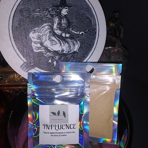 May include: A round, wooden coaster with a black and white illustration of a witch on a broom. Two iridescent pouches are in front of the coaster. One pouch has the word "INFLUENCE" printed on it.
