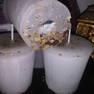 May include: Close-up of three white, cylindrical candles. The top candle is tilted, revealing a wick and embedded herbs and botanicals. The candles have a slightly textured surface and are unscented. The candles are designed for spiritual or ritual use.