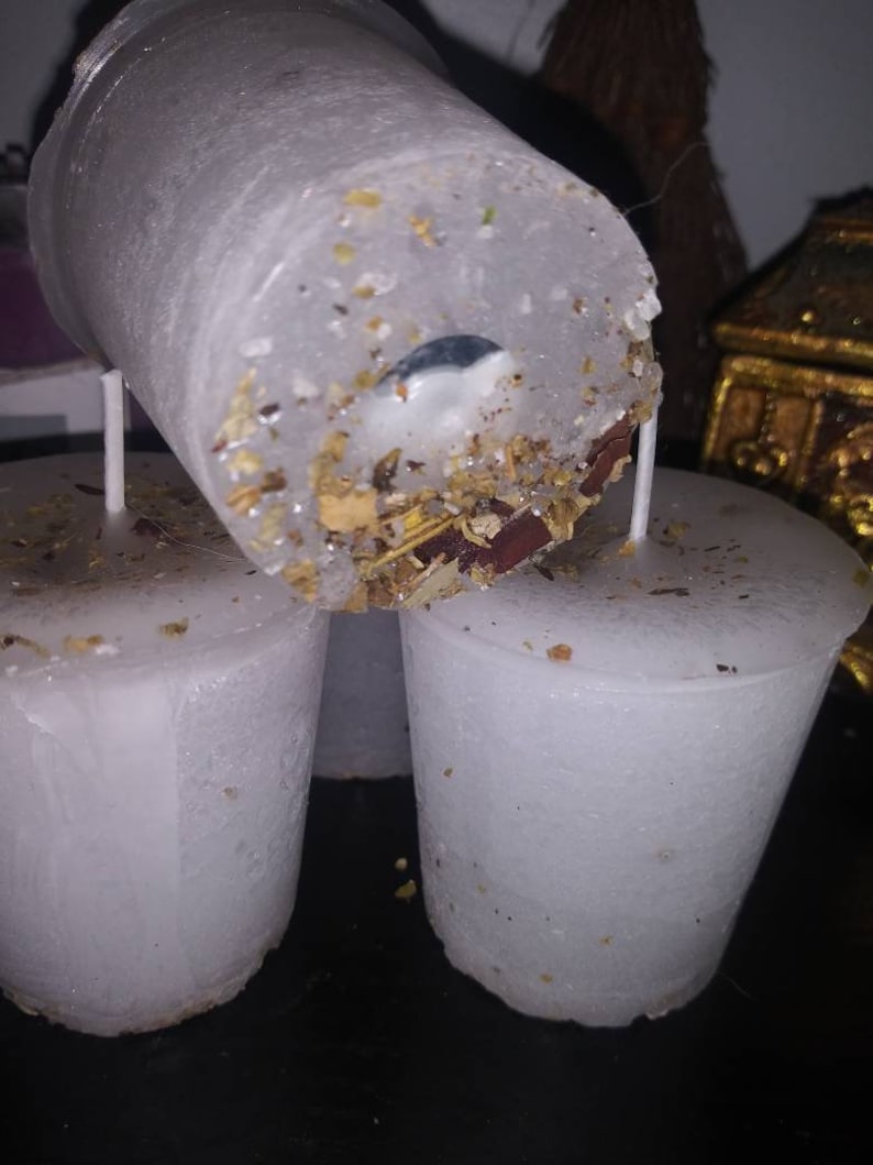 May include: Close-up of three white, cylindrical candles. The top candle is tilted, revealing a wick and embedded herbs and botanicals. The candles have a slightly textured surface and are unscented. The candles are designed for spiritual or ritual use.