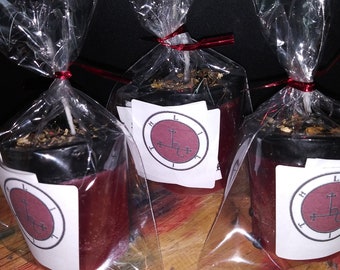 May include: Three red candles wrapped in clear cellophane, tied with red ribbon. Each candle has a white label with a circular design and the letters "L H T". The candles are on a wooden surface.