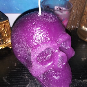 May include: A deep purple skull-shaped candle with a textured, slightly crystalline surface finish. A single white wick protrudes from the top of the cranium. The wax form features defined eye sockets, a nasal cavity, and a jawline structure.