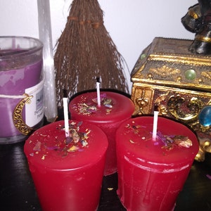 May include: Three red votive candles with white wicks are arranged in a group. The candles are decorated with dried flowers and herbs. A small broom, a purple candle, and a decorative box are in the background.