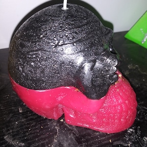 May include: A black and red skull-shaped candle with a wick. The top half of the candle is black, while the bottom half is red. The candle has a textured surface.