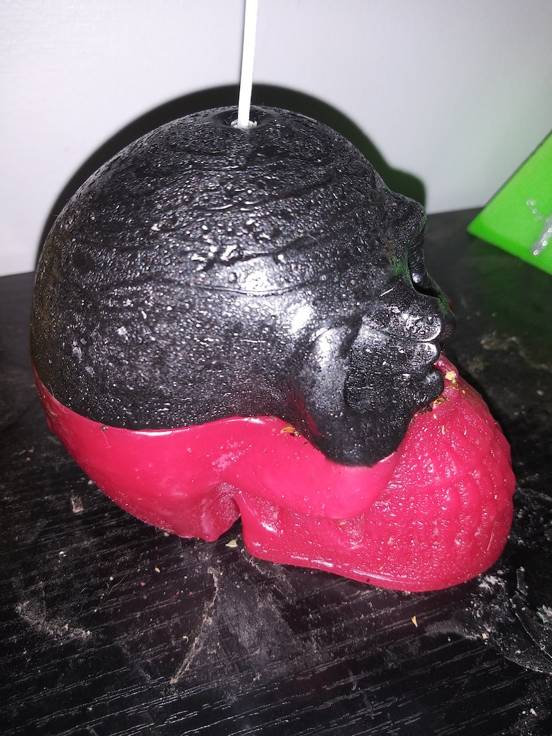 May include: A black and red skull-shaped candle with a wick. The top half of the candle is black, while the bottom half is red. The candle has a textured surface.
