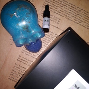 May include: A teal skull-shaped candle with a purple sphere in its mouth, a small bottle labeled "Speak Now," and a black box with a sticker. The background features handwritten instructions on aged paper.