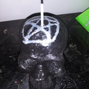 May include: A black skull-shaped candle with a white pentagram design on top. A white wick extends from the center of the skull. The candle has a glossy finish and sits on a dark surface.
