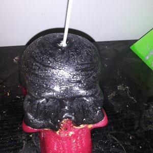 May include: A skull-shaped candle with a black head and a red tongue. The candle has a white wick and is placed on a dark surface. The skull's head has a textured appearance.