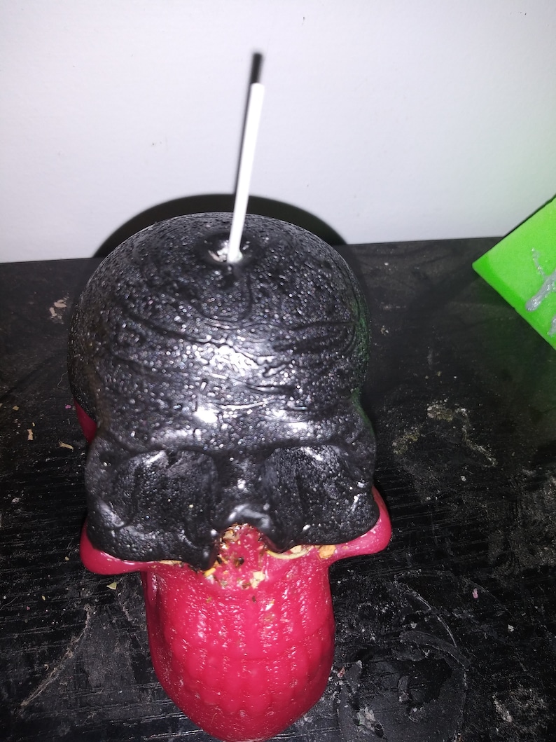 May include: A skull-shaped candle with a black head and a red tongue. The candle has a white wick and is placed on a dark surface. The skull's head has a textured appearance.