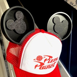 Puede incluir: Gorra de camionero roja y blanca con el logotipo de Pizza Planet, combinada con orejas de Mickey Mouse negras. La gorra tiene una visera roja y una parte trasera de malla, con el logotipo de Pizza Planet en rojo. Las orejas de Mickey Mouse son negras con textura brillante.