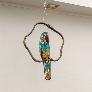 May include: A mosaic parrot sculpture hanging from a metal ring. The parrot is blue, yellow, orange, and red with black eyes. The parrot is perched on a branch.