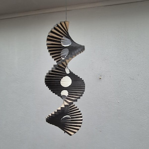 May include: A hanging wooden sculpture with a spiral design. The sculpture features alternating black and natural wood-toned segments, with white and grey accents. The design includes circular and curved shapes, suspended from a thin wire.