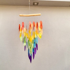 May include: A rainbow wind chime made of paper leaves hanging from a wooden branch. The leaves are arranged in a teardrop shape and are suspended from a string.