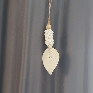 May include: A white and beige tassel with a leaf-shaped design hangs from a grey curtain. The tassel is made of small white beads and a woven leaf.