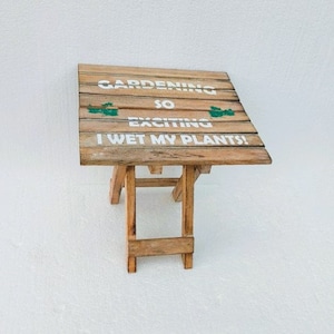 May include: A wooden folding table with a painted sign that reads "Gardening so exciting I wet my plants!" The table is made of natural wood with a light brown finish.