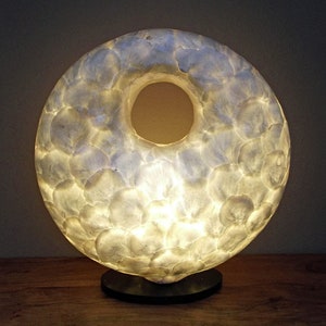 May include: A distinctive, illuminated lamp in a circular form, resembling a donut. The lamp is adorned with a white, textured material, possibly capiz shells, and radiates a warm light. It rests on a small, round black base, offering a striking contrast.