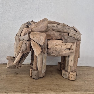 Pu&ograve; includere: Una scultura di elefante in legno fatta a mano. L'elefante &egrave; costruito con vari pezzi di legno di deriva, mostrando un'estetica naturale e rustica. La scultura &egrave; esposta su una superficie di legno contro un muro bianco.