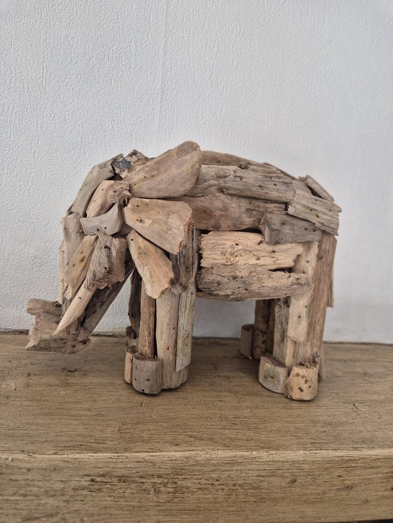 Pu&ograve; includere: Una scultura di elefante in legno fatta a mano. L'elefante &egrave; costruito con vari pezzi di legno di deriva, mostrando un'estetica naturale e rustica. La scultura &egrave; esposta su una superficie di legno contro un muro bianco.