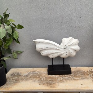 May include: White wooden sculpture of a seashell on a black stand. The sculpture is on a light wood table with carved floral designs.