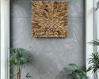 May include: A square, wooden wall art piece with a sunburst design made of small, light brown wooden pieces. The artwork is mounted on a gray stone wall, with potted plants in the foreground.