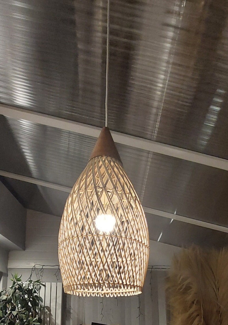 May include: A woven wicker pendant light fixture with a teardrop shape hangs from a white cord. The light fixture is made of natural materials and has a warm, inviting aesthetic.