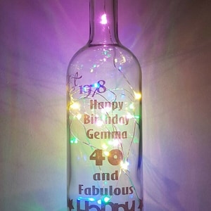 May include: A clear glass bottle with a cork top and fairy lights inside. The bottle has a gold vinyl design that says "Happy Birthday Gemma 40 and Fabulous Happy Birthday".