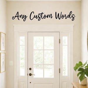 Może przedstawiać: Jasne, dobrze oświetlone wejście z białymi drzwiami i naświetlami. Nad drzwiami napisano „Any Custom Words” stylowym pismem. Drewniany stolik konsolowy mieści roślinę doniczkową.