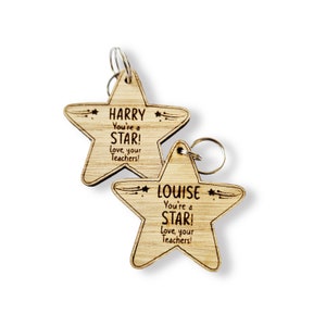 Może przedstawiać: Dwa drewniane breloki w kształcie gwiazdy z tekstem "You're a STAR! Love, your Teachers!" i wygrawerowanymi imionami "HARRY" i "LOUISE".