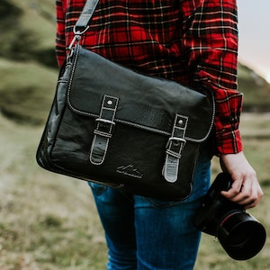 Könnte beinhalten: Schwarze Leder-Umhängetasche mit weißen Nähten und silbernen Schnallen. Die Tasche hat einen Klappverschluss und einen Schulterriemen. Eine Kamera wird in der Hand gehalten. Die Person trägt ein rot-schwarz kariertes Hemd und blaue Jeans.