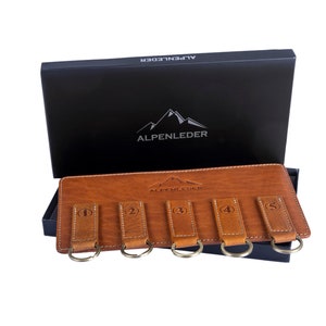 Könnte beinhalten: Ein brauner Leder-Schlüsselorganisator mit fünf Schlüsselringen. Jeder Ring hat ein nummeriertes Leder-Etikett. Der Organisator ist geöffnet und die Box, in der er geliefert wurde, befindet sich dahinter. Die Box ist schwarz mit dem Firmenlogo "ALPENLEDER", das in Weiß gedruckt ist.