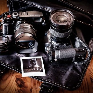 Może przedstawiać: Dwa czarne aparaty fotograficzne z obiektywami i czarna sk&oacute;rzana torba na aparat na drewnianej powierzchni.