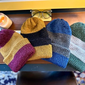 May include: Four striped knit hats in different colors: burgundy and gold, black and gold, gray and blue, and gray and green.