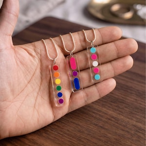 May include: Three colorful pendant necklaces. Each necklace features a clear, rectangular pendant with a silver chain. One pendant has a rainbow of dots, another has a pink rectangle and blue dot, and the last has pink, white, and blue dots.