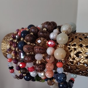 May include: A stack of colorful beaded bracelets with various textures and materials, including wood, glass, and stone. The bracelets are arranged on a gold metal stand.