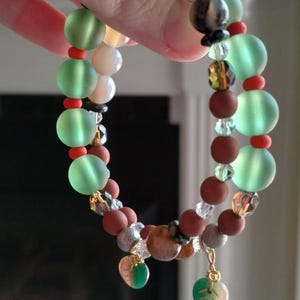 May include: A beaded bracelet featuring a variety of colorful beads. The bracelet includes large, frosted green beads, small red beads, and brown, white, and clear beads. Two leaf-shaped charms dangle from the bracelet.