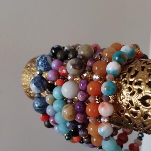 May include: A stack of colorful beaded bracelets with various stones and textures, including turquoise, orange, red, purple, and white beads. The bracelets are displayed on a gold metal stand.
