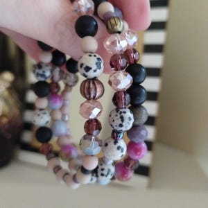 May include: A set of three beaded bracelets with a variety of beads in shades of black, pink, purple, and cream. The bracelets feature round, faceted, and patterned beads.