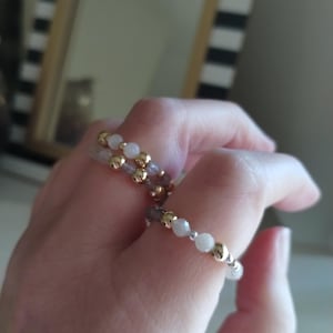 May include: Two beaded rings with gold and white beads. The rings are made of small, round beads strung together. The gold beads are interspersed with white and gray beads. The rings are on a person's finger.