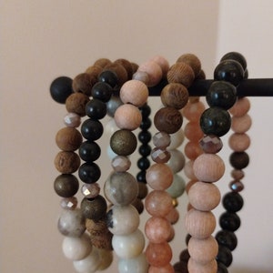 May include: A set of six beaded bracelets in various colors and materials, including wood, stone, and metal. The bracelets are stacked on a black stand.