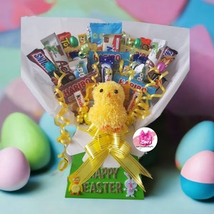 Puede incluir: Un peluche de pollito amarillo se sienta en una cesta de regalo blanca llena de barras de chocolate y dulces surtidos. La cesta está decorada con una cinta amarilla y un letrero verde que dice "Feliz Pascua".