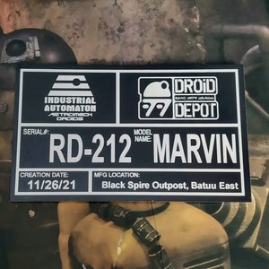 Puede incluir: Una placa negra y plateada con el texto "RD-212 MARVIN" y los logotipos "DROID DEPOT". La placa incluye la fecha de creaci&oacute;n "11/26/21" y el lugar de fabricaci&oacute;n "Black Spire Outpost, Batuu East".