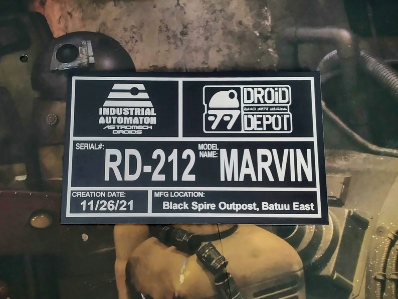 Puede incluir: Una placa negra y plateada con el texto "RD-212 MARVIN" y los logotipos "DROID DEPOT". La placa incluye la fecha de creaci&oacute;n "11/26/21" y el lugar de fabricaci&oacute;n "Black Spire Outpost, Batuu East".