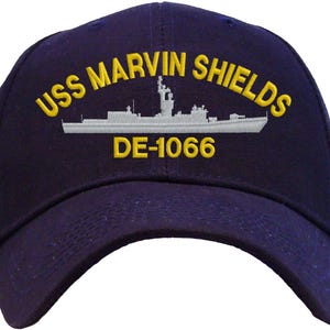 May include: Navy blue baseball cap with the embroidered text "USS MARVIN SHIELDS" in yellow, along with a ship illustration and the number "DE-1066". The cap has a curved brim and a structured crown.
