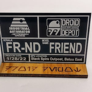 Puede incluir: Letrero negro y plateado con el texto "FR-ND FRIEND" y los logotipos "DROID DEPOT". El letrero incluye el texto "INDUSTRIAL AUTOMATON ASTROMECH DROIDS" y "1/28/22". El letrero est&aacute; montado sobre una base de madera.