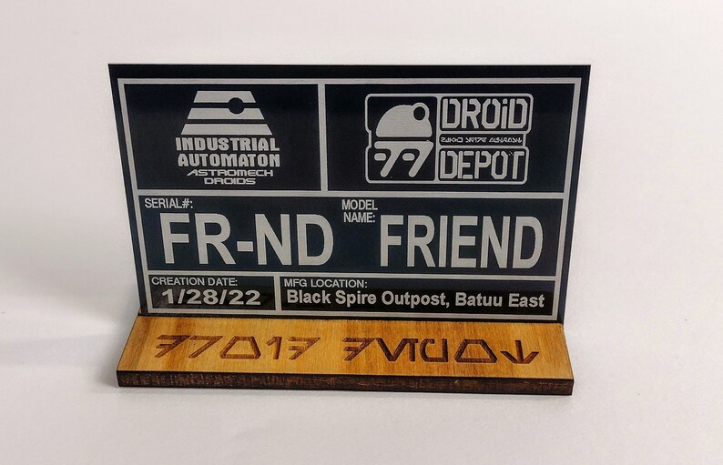 Puede incluir: Letrero negro y plateado con el texto "FR-ND FRIEND" y los logotipos "DROID DEPOT". El letrero incluye el texto "INDUSTRIAL AUTOMATON ASTROMECH DROIDS" y "1/28/22". El letrero est&aacute; montado sobre una base de madera.