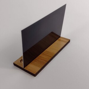 Puede incluir: Un soporte de acr&iacute;lico gris oscuro para letreros o fotos, exhibido sobre una base de madera. La base es rectangular con un acabado de veta de madera natural. El letrero est&aacute; en &aacute;ngulo, creando una exhibici&oacute;n moderna.