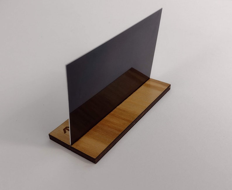 Puede incluir: Un soporte de acr&iacute;lico gris oscuro para letreros o fotos, exhibido sobre una base de madera. La base es rectangular con un acabado de veta de madera natural. El letrero est&aacute; en &aacute;ngulo, creando una exhibici&oacute;n moderna.