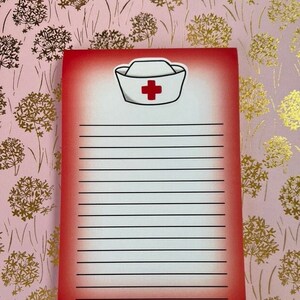 May include: Red notepad with a white nurse's cap and a red cross on a pink background with gold floral pattern.