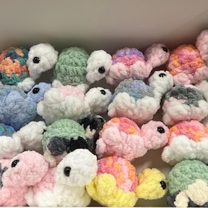 May include: A collection of crocheted turtle toys in various colors, including pink, blue, green, and white. The turtles have black eyes and are made with a soft, fluffy yarn.