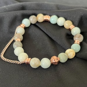 May include: A green and brown beaded bracelet with a rose gold clasp and a rose gold charm. The bracelet is made of natural stones and is designed to be worn on the wrist.