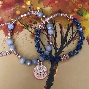 May include: Two bracelets with a silver and blue color scheme. One bracelet has a silver charm with a sunburst design. The bracelets are on a background with a watercolor painting of a tree.